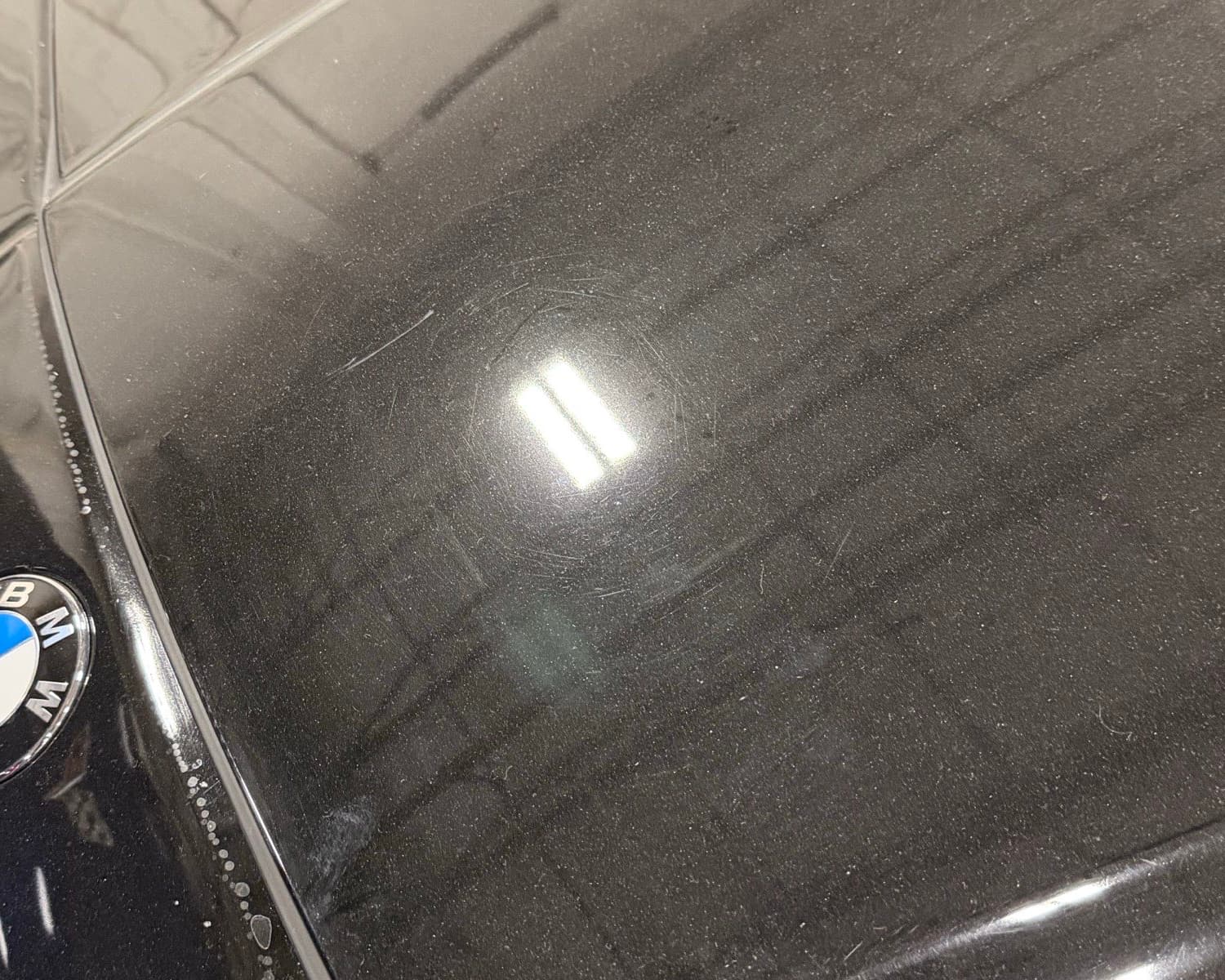 The rear deck of a black BMW, where a halo of fine circular scratches surrounds the reflection of a strip light, with dust across the panel and dried water spots gathered along its lower edge.
